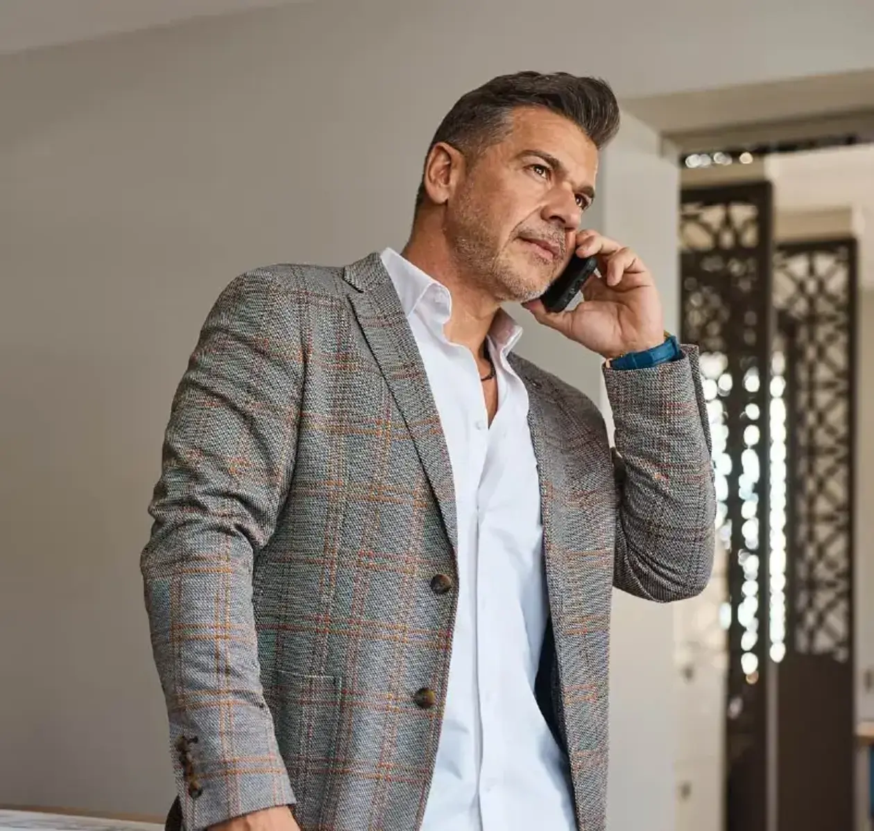 A man in a grey tweed blazer and white dress shirt takes a phone call.