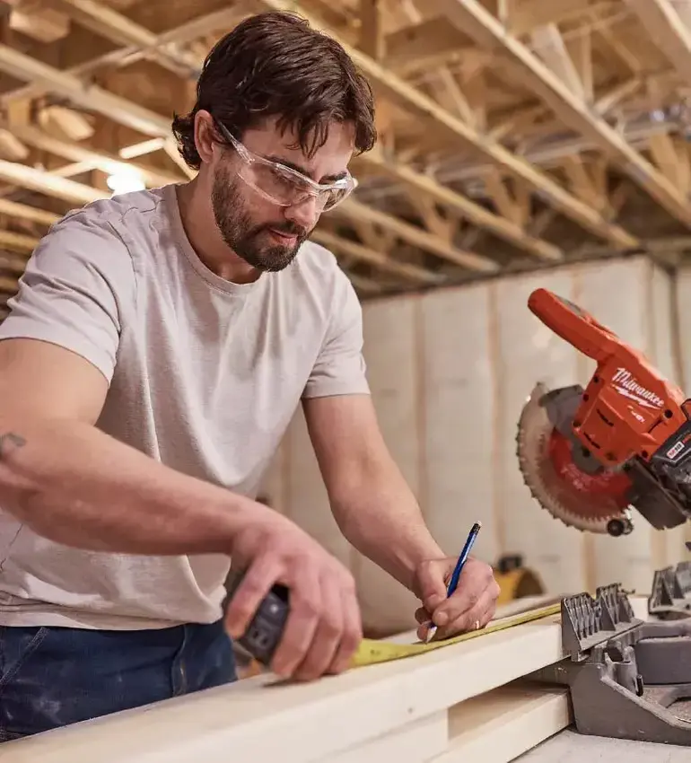 A man wearing safety glasses takes measurements on a piece of lumber next to a buzzsaw.