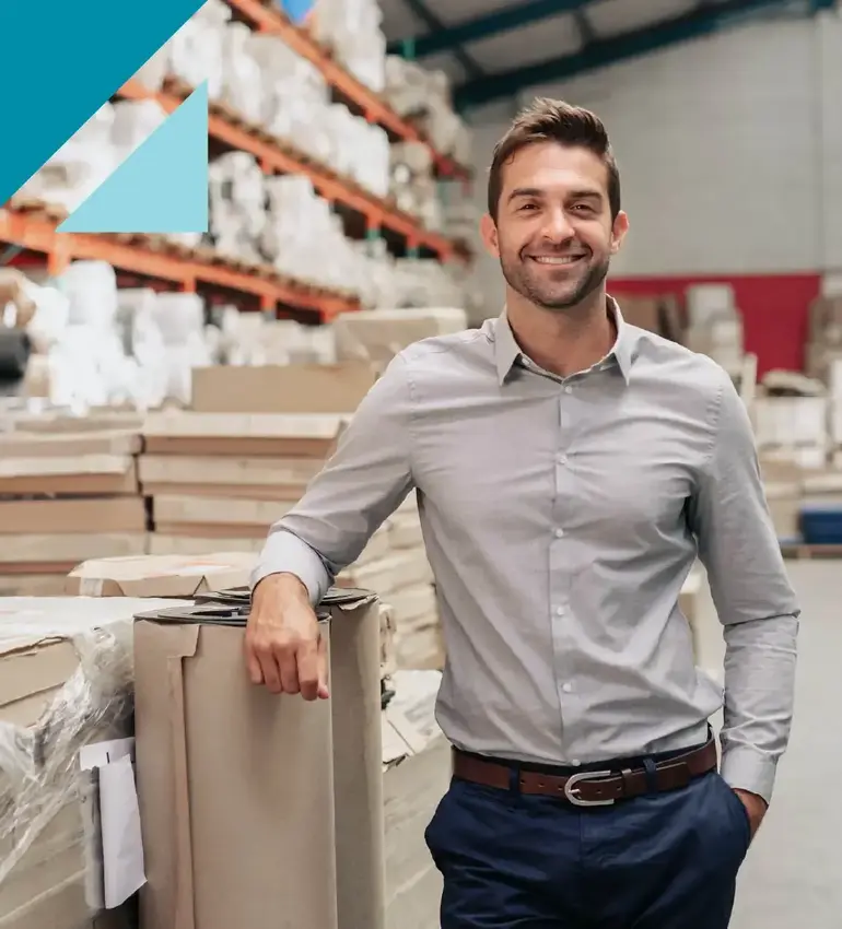 A young man wearing a grey dress shirt smiles at the camera in a warehouse.
