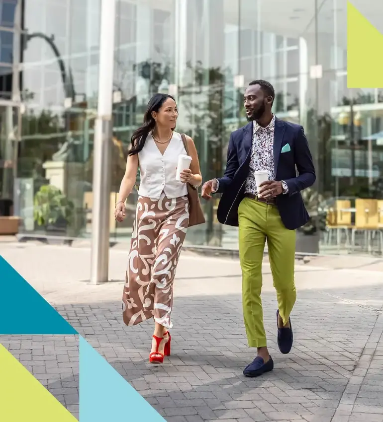 A man and woman in modern dress attire engage in conversation as they stroll through an urban landscape.