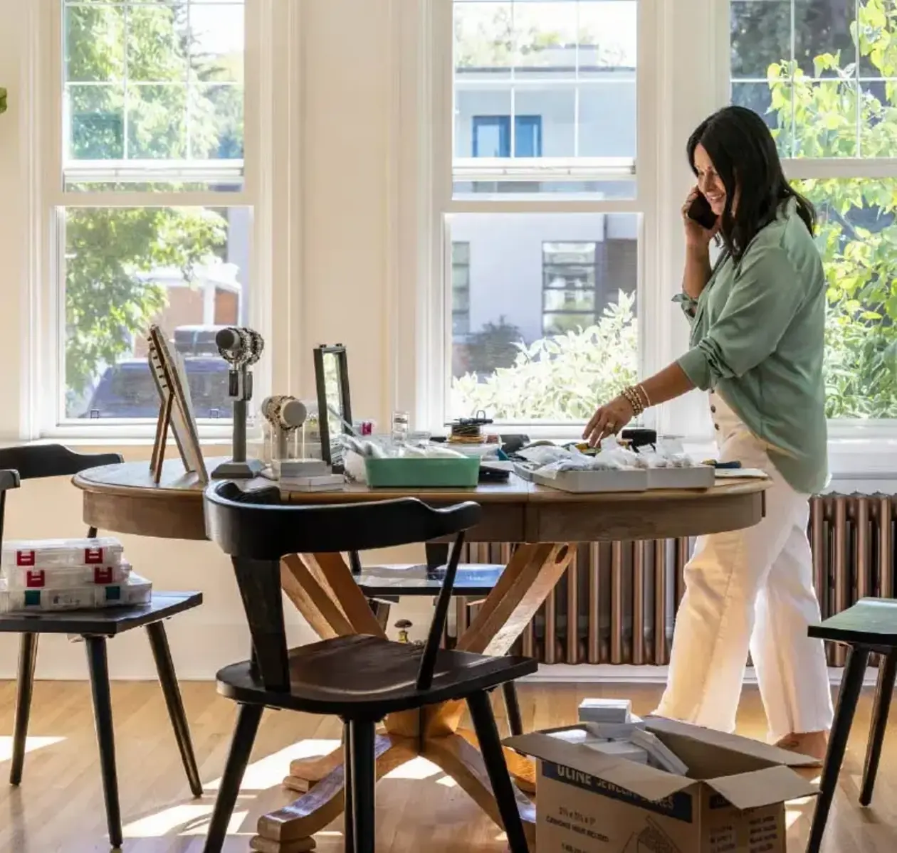A woman in comfortable clothing chats on the phone while taking stock of items for sale on a kitchen table.