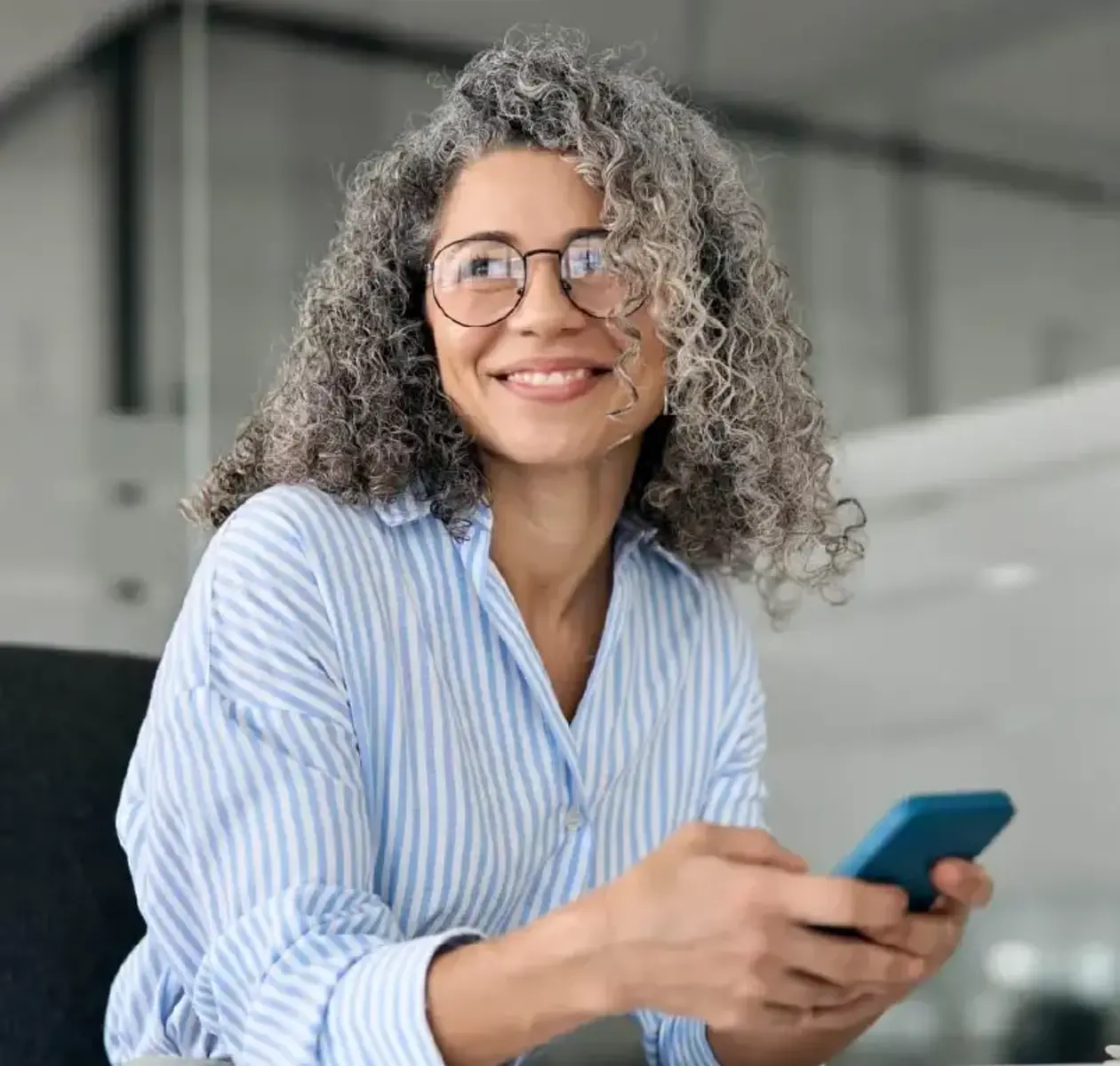A woman with glasses and striped dress shirt holds her phone and smiles off camera.