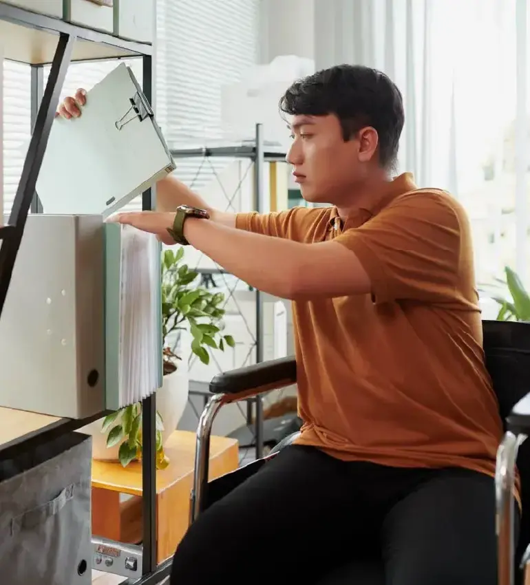 A young man seated in a bright living room organizes his work shelf.