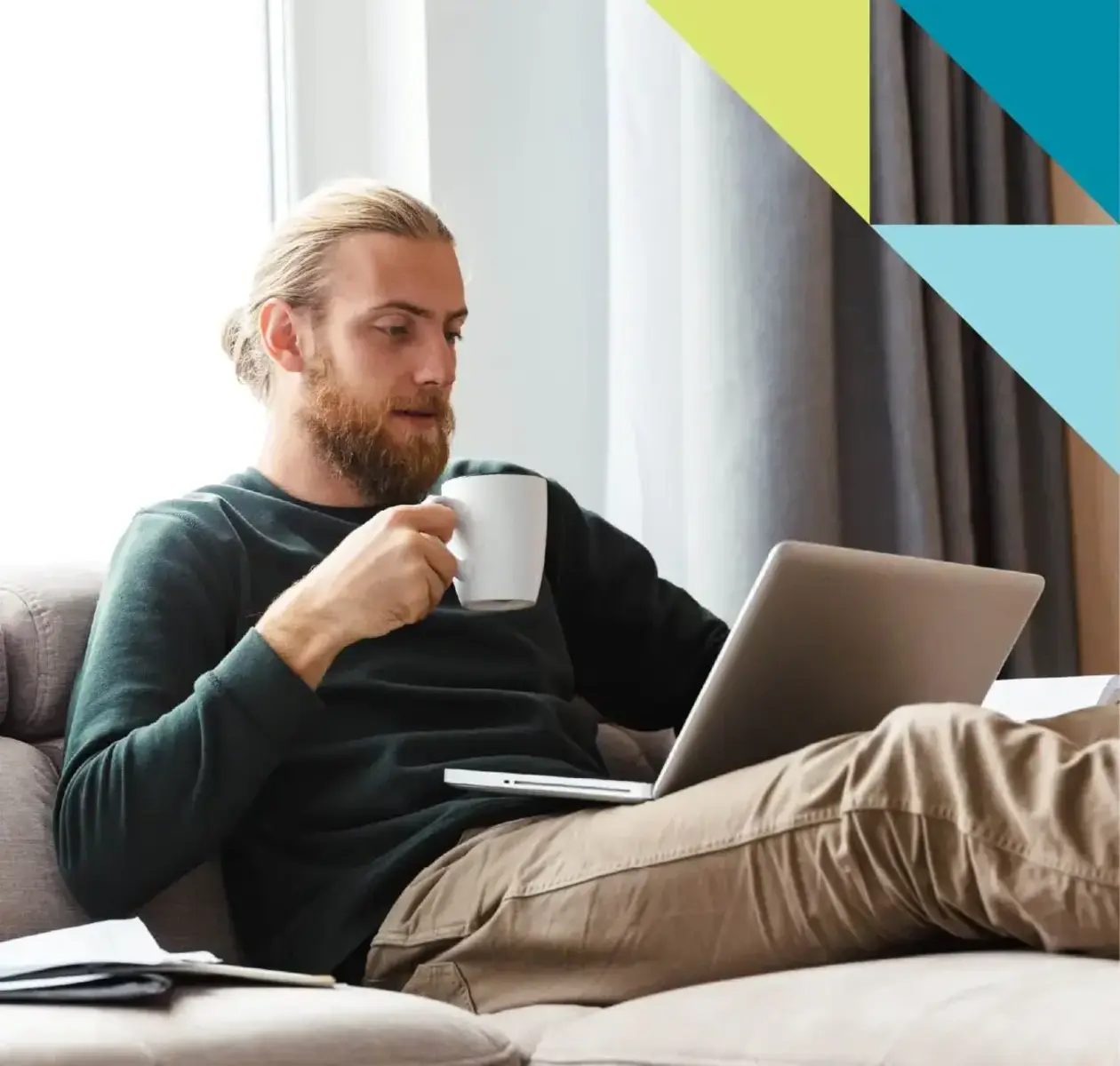 A young man with blonde hair and beard sips a cup of coffee while stretched out on the couch with his laptop.