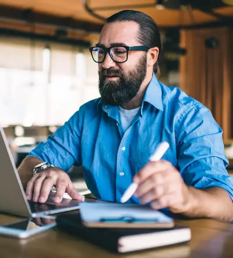 A bearded man with glasses in a blue button-up shirt completes some work on his laptop computer.