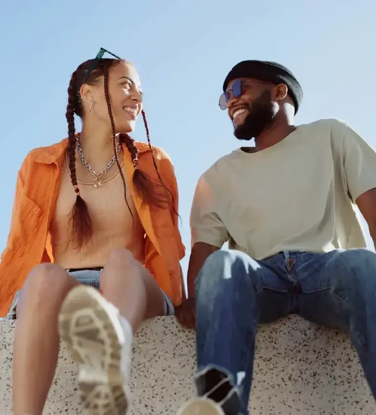 A woman in an orange shirt and pigtails smiles at a man next to her who’s wearing sunglasses and a toque.
