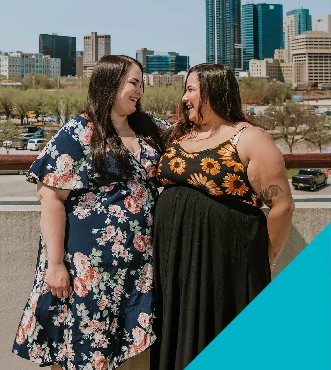 Two young women in flower-print sundresses look at each other and smile.