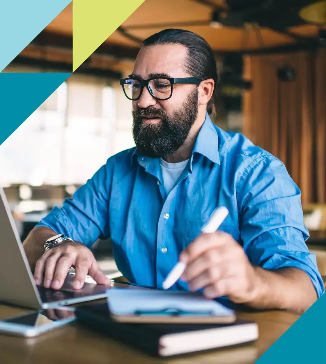 A bearded man with glasses in a blue button-up shirt completes some work on his laptop computer.