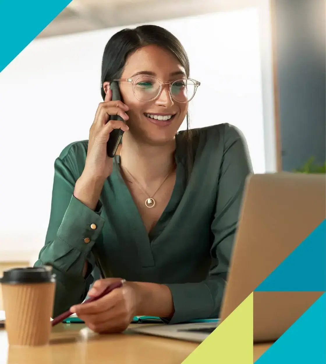 A young woman wearing glasses and a green blouse takes a call while keeping an eye on her laptop.