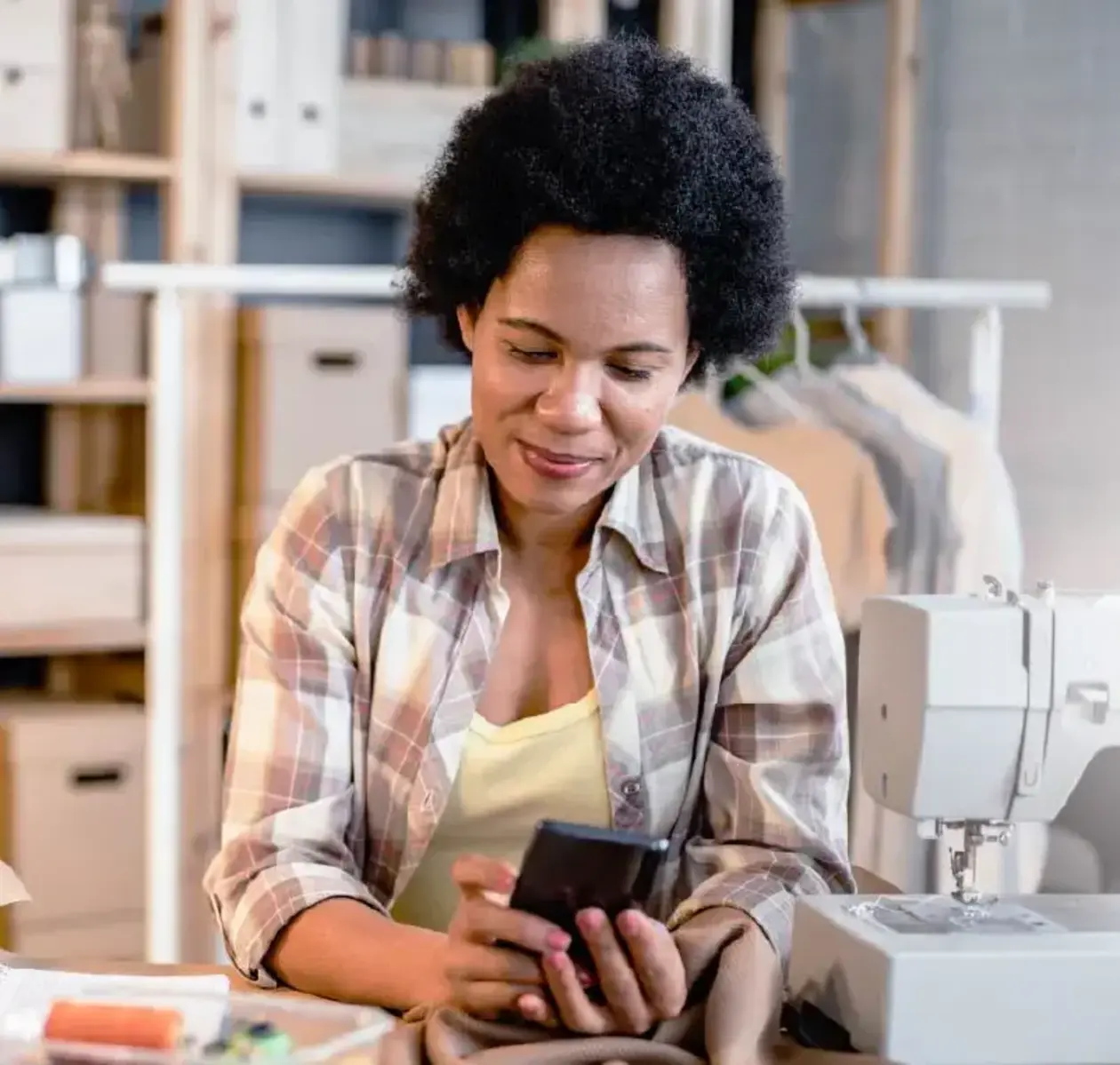A woman in a plaid work shirt surrounded by sewing equipment and materials checks her phone. 