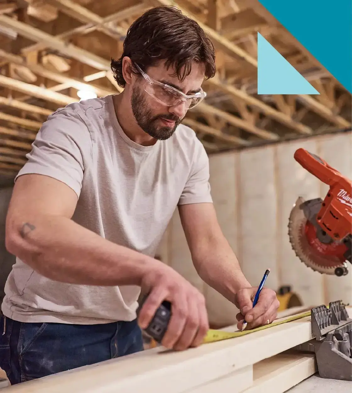 A man wearing safety glasses takes measurements on a piece of lumber next to a buzzsaw.