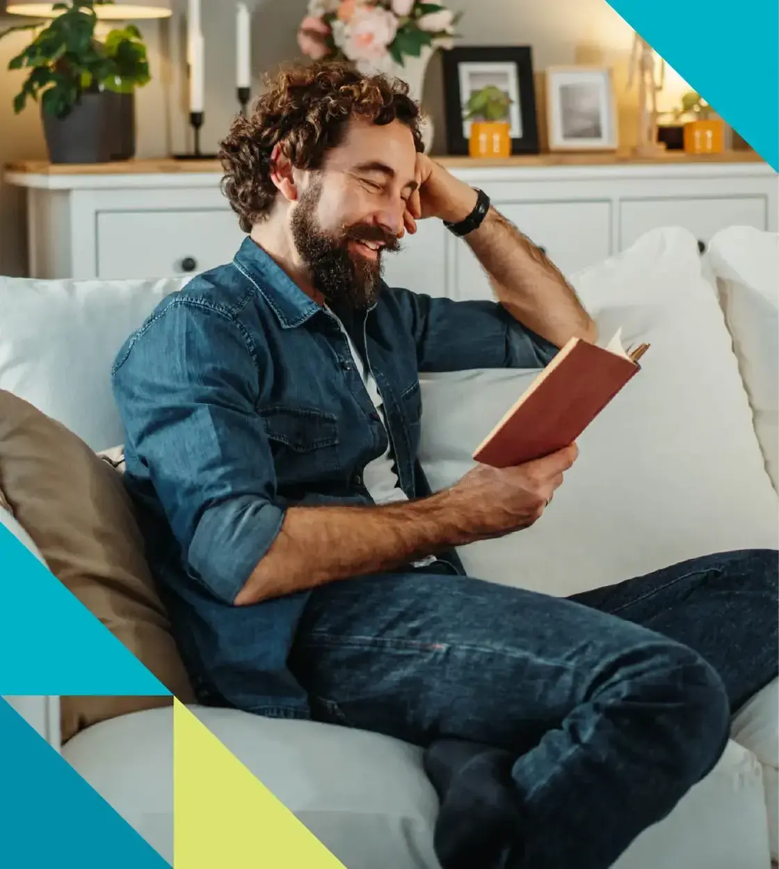A bearded man clad in denim cozies up on the couch to read a good book.