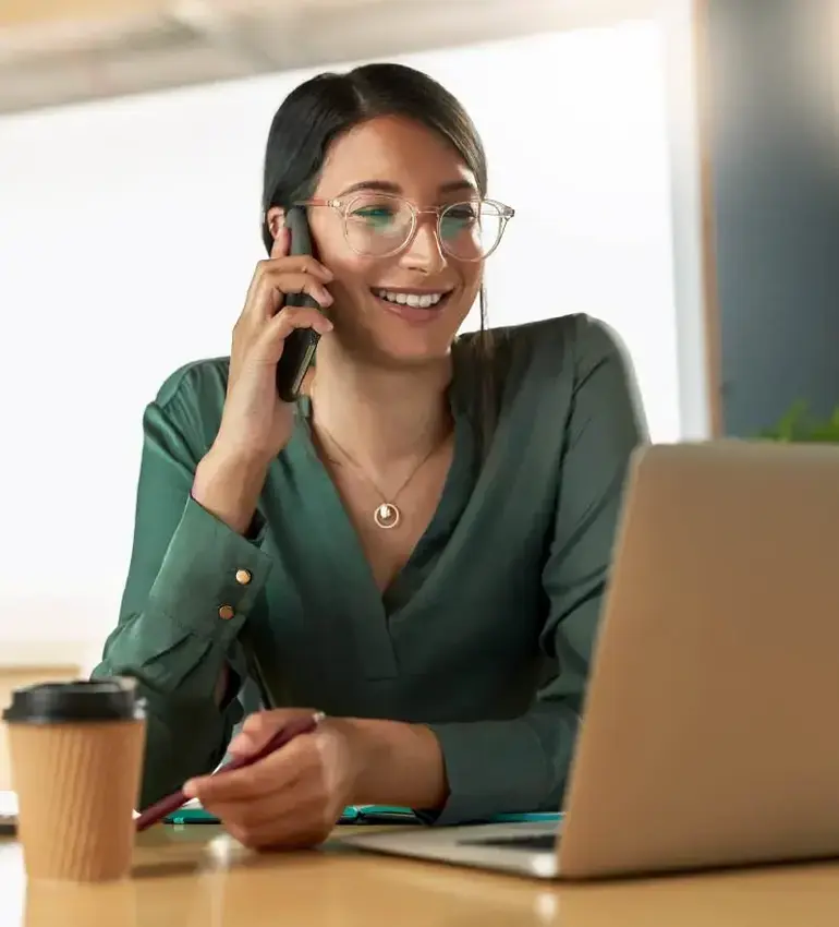 A young woman wearing glasses and a green blouse takes a call while keeping an eye on her laptop.