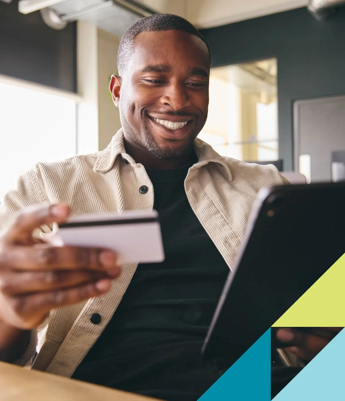 A man wearing a cream corduroy overshirt smiles while checking his bank balance on a tablet.