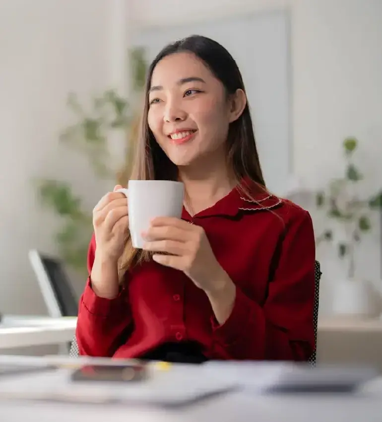 A young woman in a red blouse smiles off camera while holding a cup of coffee.