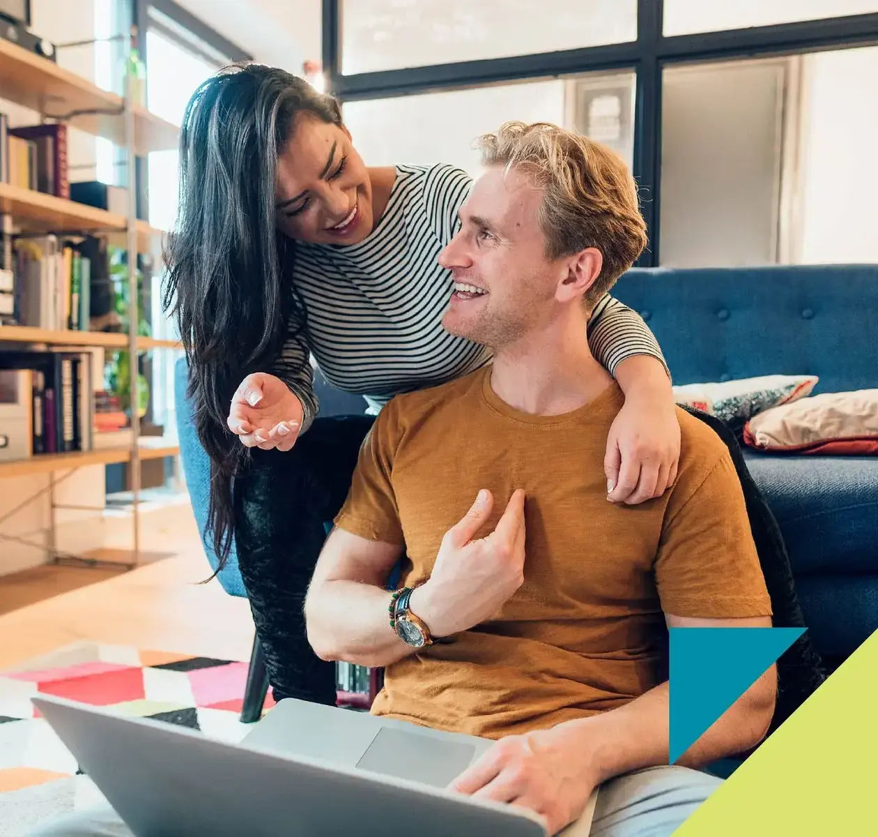 A happy young couple have fun using a laptop in their brightly lit living room.