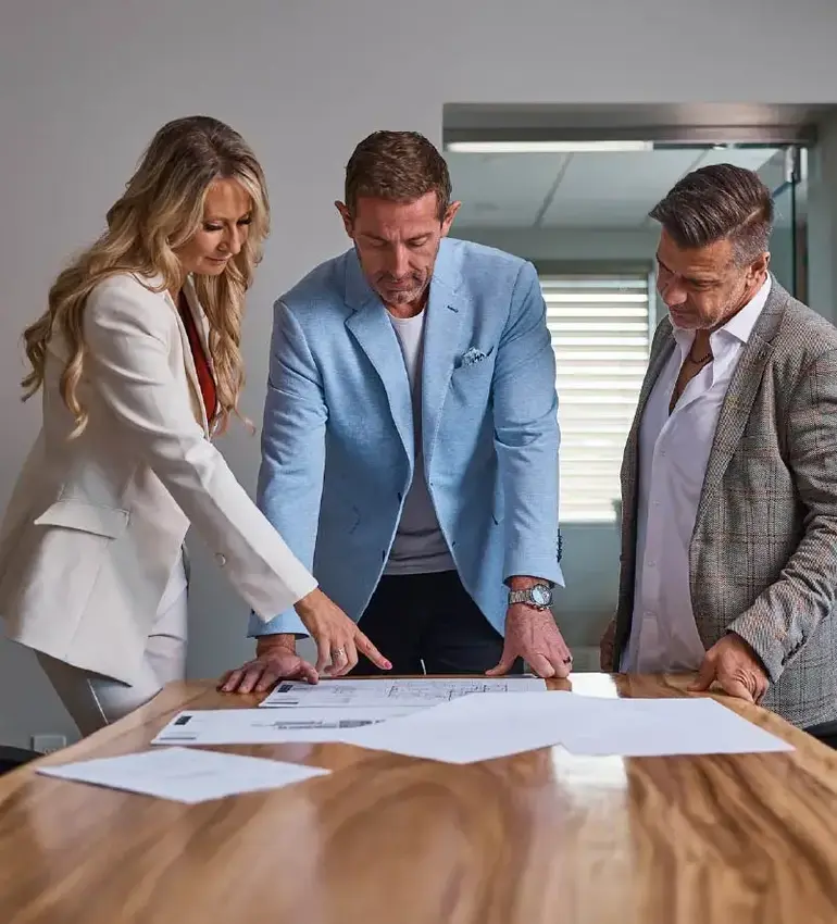 Three business professionals examine blueprints on a large table.