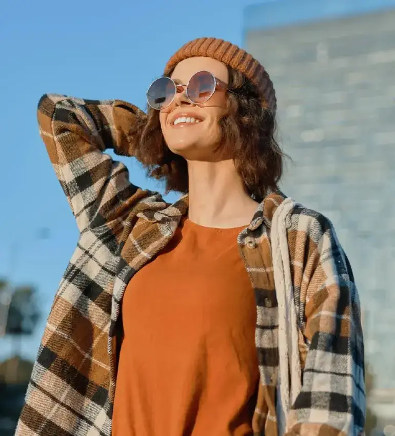 A young woman wearing flannel, sunglasses and a toque smiles into the autumn sun.