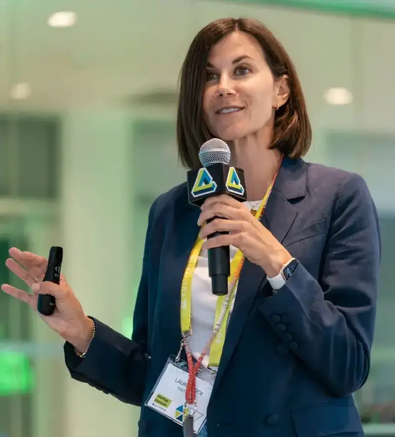 A woman with medium-length brown hair wearing a blue blazer speaks into a microphone.
