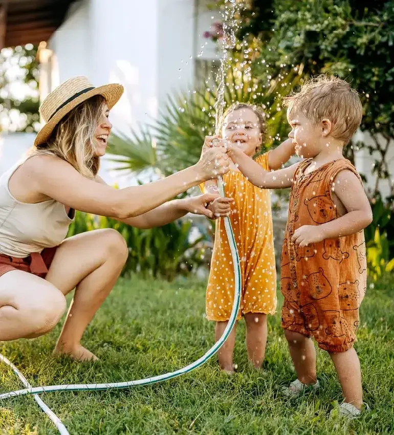 A mom and her two toddlers soak up some summer fun while playing with a garden hose in the yard.