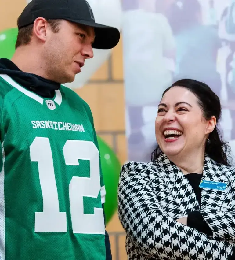 A female employee in a black and white checkered blazer laughs with a man in a Roughriders football jersey.