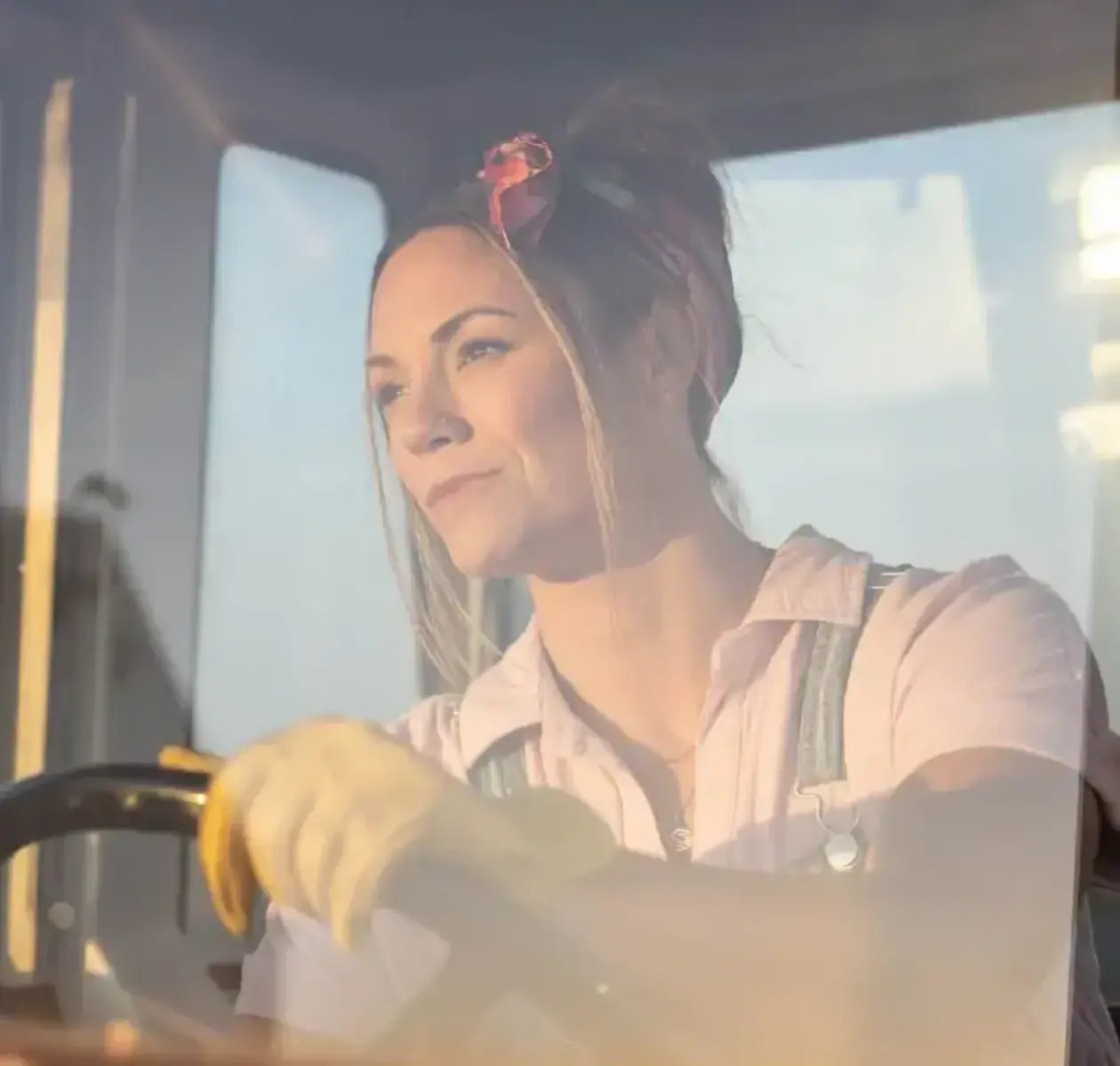A female farmer operates a piece of heavy equipment as sunshine beams into the cab.