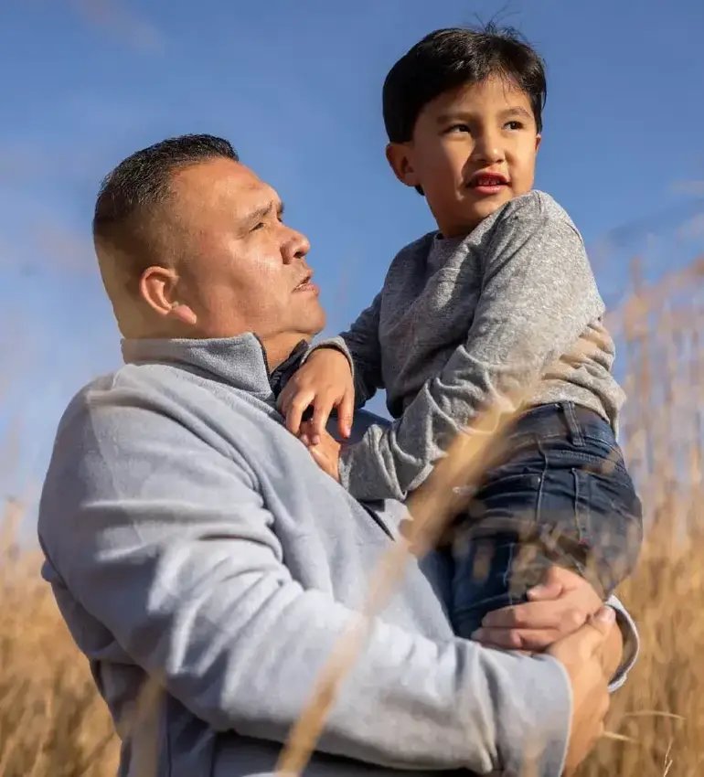 A father carries his young son through a wheat field on a clear blue day.