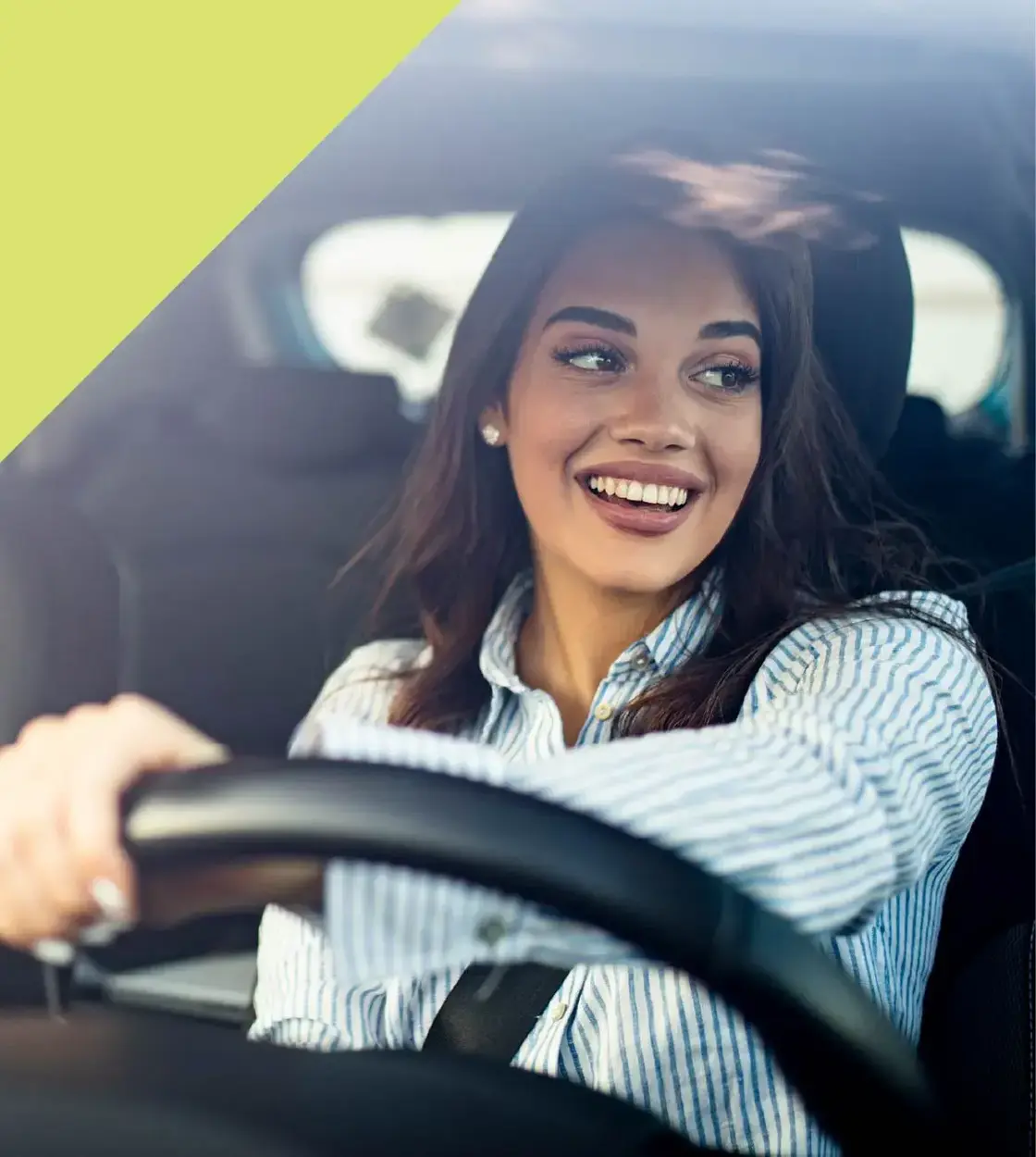 A woman wearing a striped dress shirt smiles out the window as she steers her vehicle along the road.
