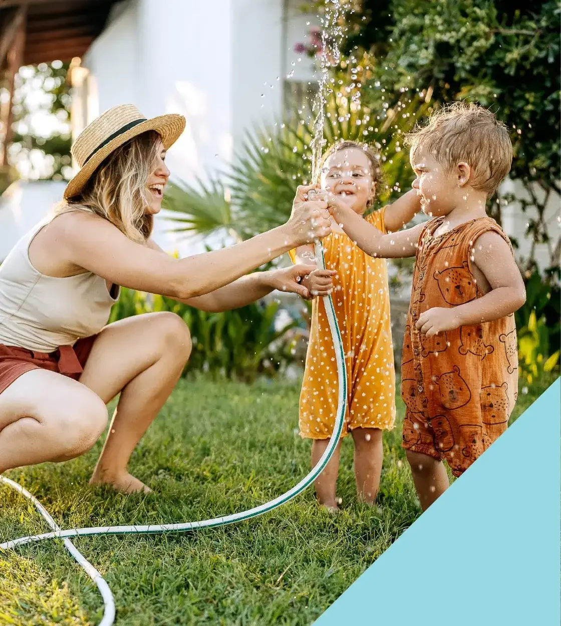 A mom and her two toddlers soak up some summer fun while playing with a garden hose in the yard.