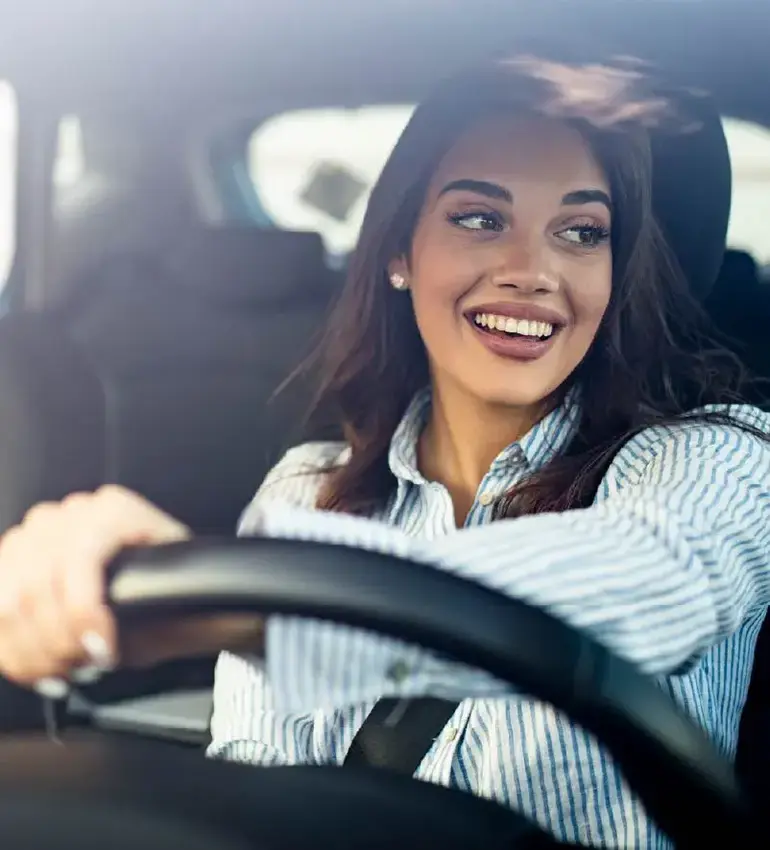 A woman wearing a striped dress shirt smiles out the window as she steers her vehicle along the road.