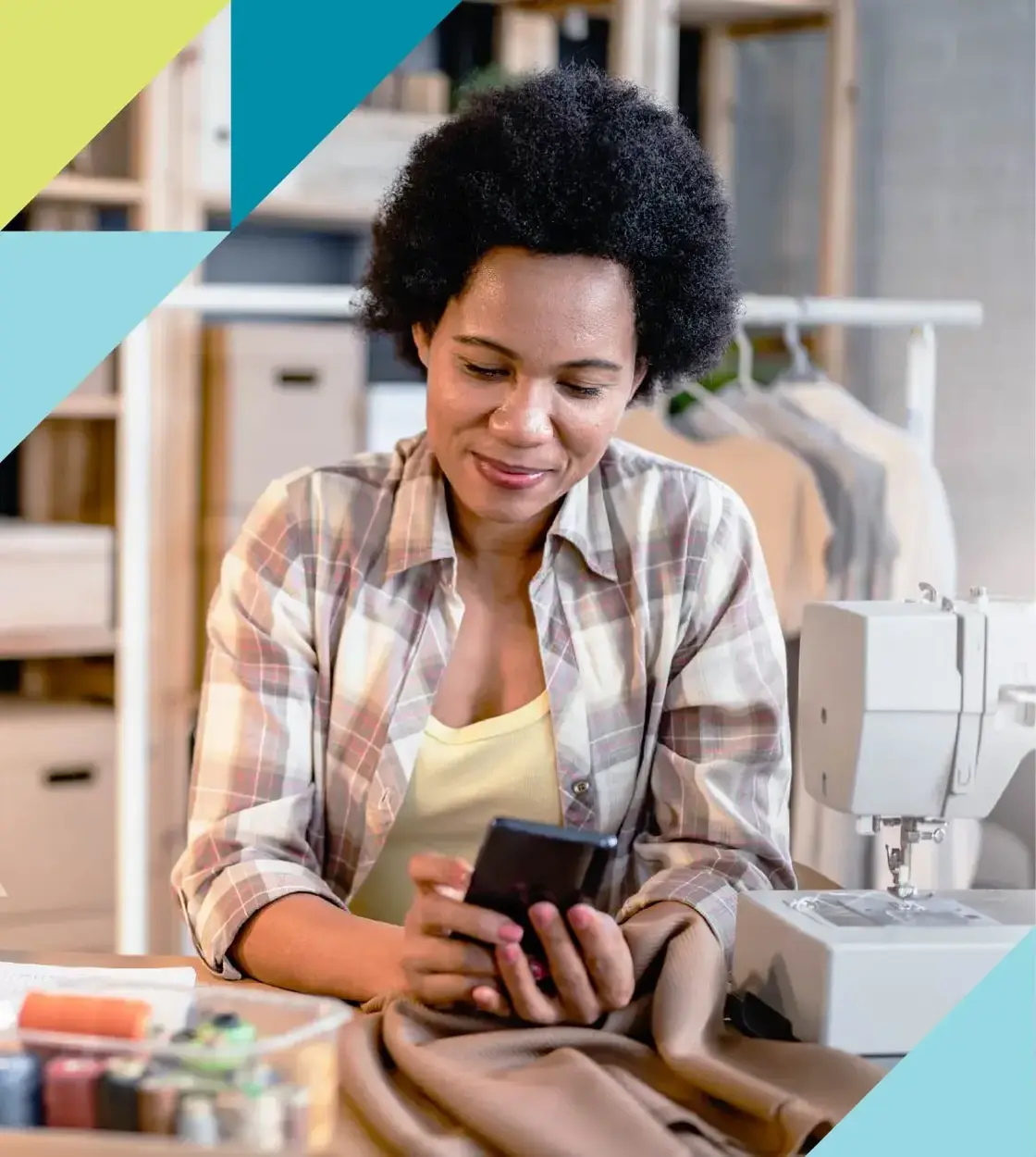 A woman in a plaid work shirt surrounded by sewing equipment and materials checks her phone. 