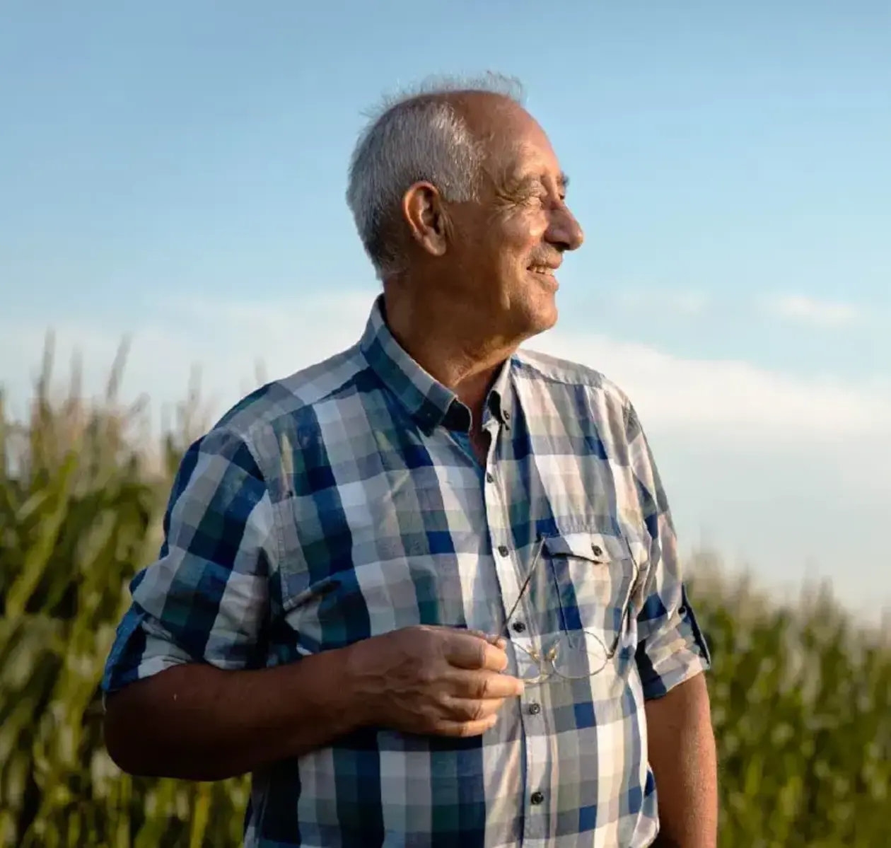 A man in a plaid shirt holds his glasses as he looks off into the distance with a contented smile.