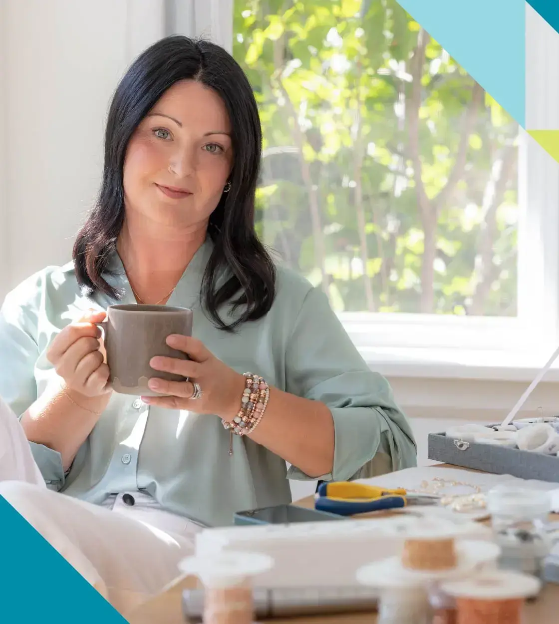 A seated woman with long dark hair holding a cup of coffee looks into the camera.