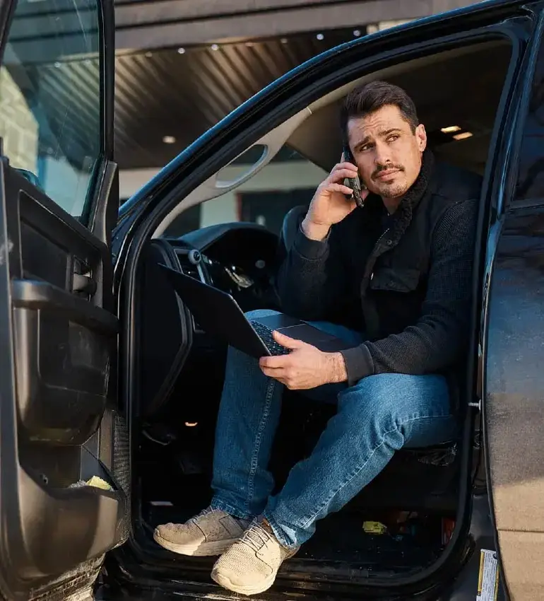 A man with dark hair and goatee leans out his vehicle while taking a call and holding his laptop computer.