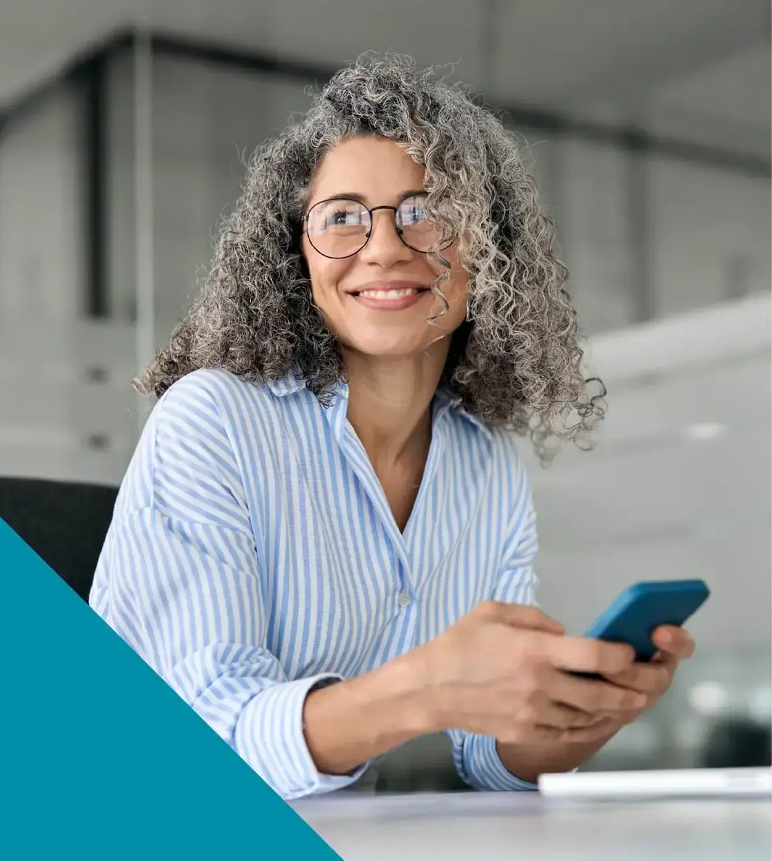 A woman with glasses and striped dress shirt holds her phone and smiles off camera.