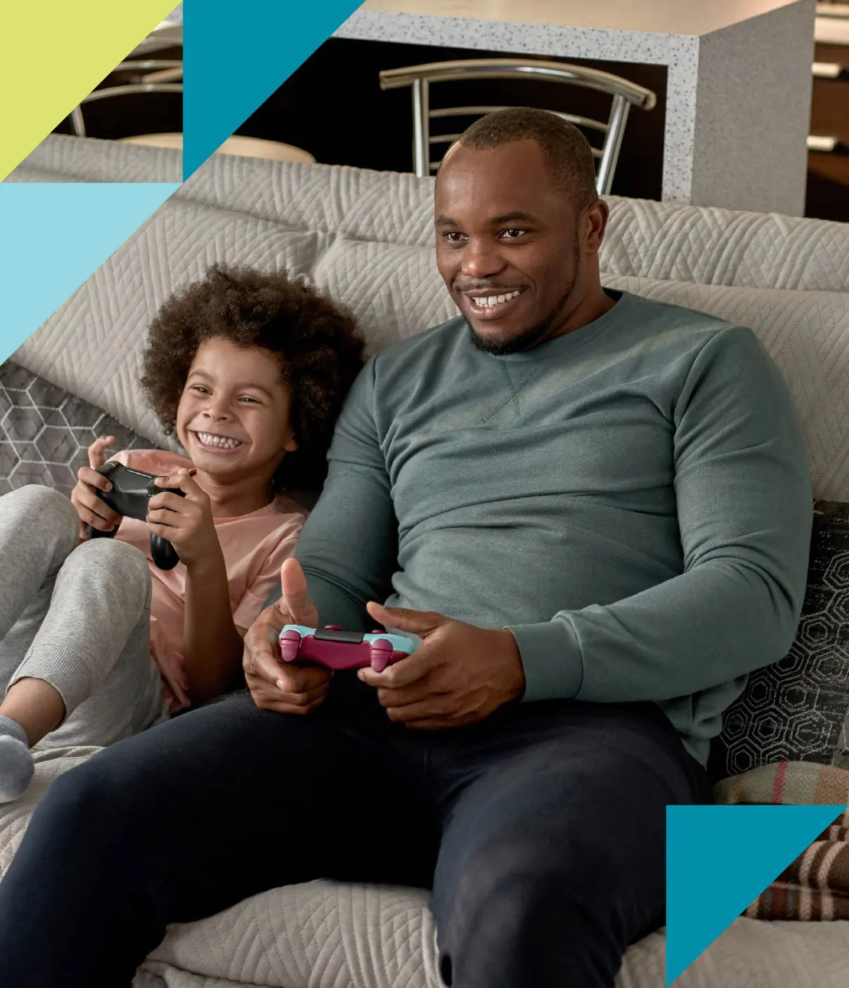 A father and child in comfy loungewear laugh as they play a gaming console on the couch.