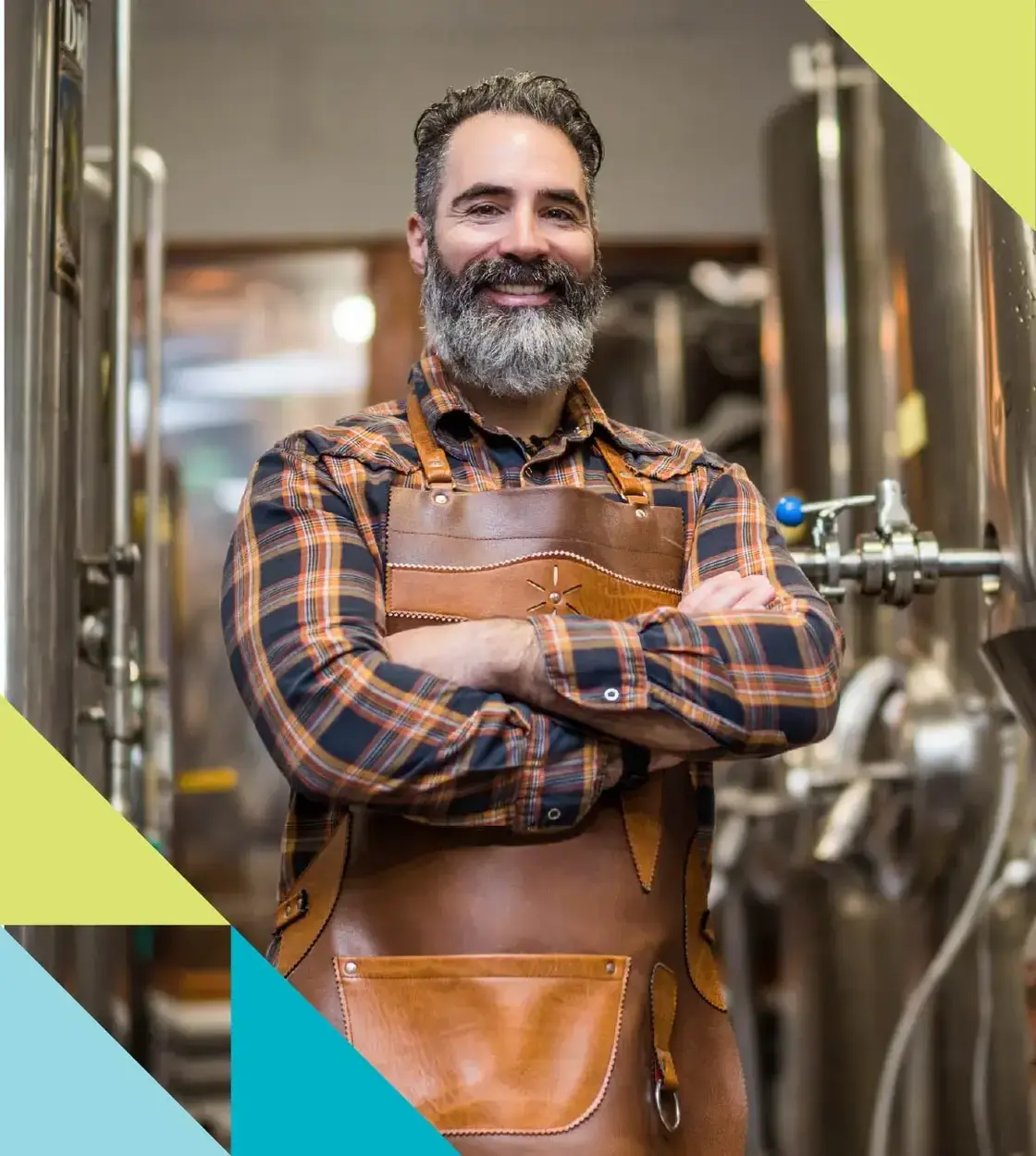 A man with a grey beard wearing a leather apron smiles at the camera while surrounded by microbrewery equipment.