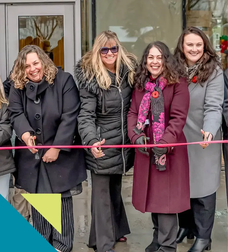Four women dressed in winter attire smile while cutting a red ribbon. 