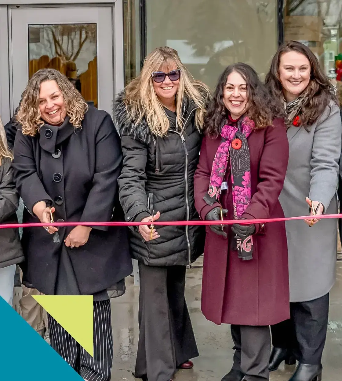 Four women dressed in winter attire smile while cutting a red ribbon. 