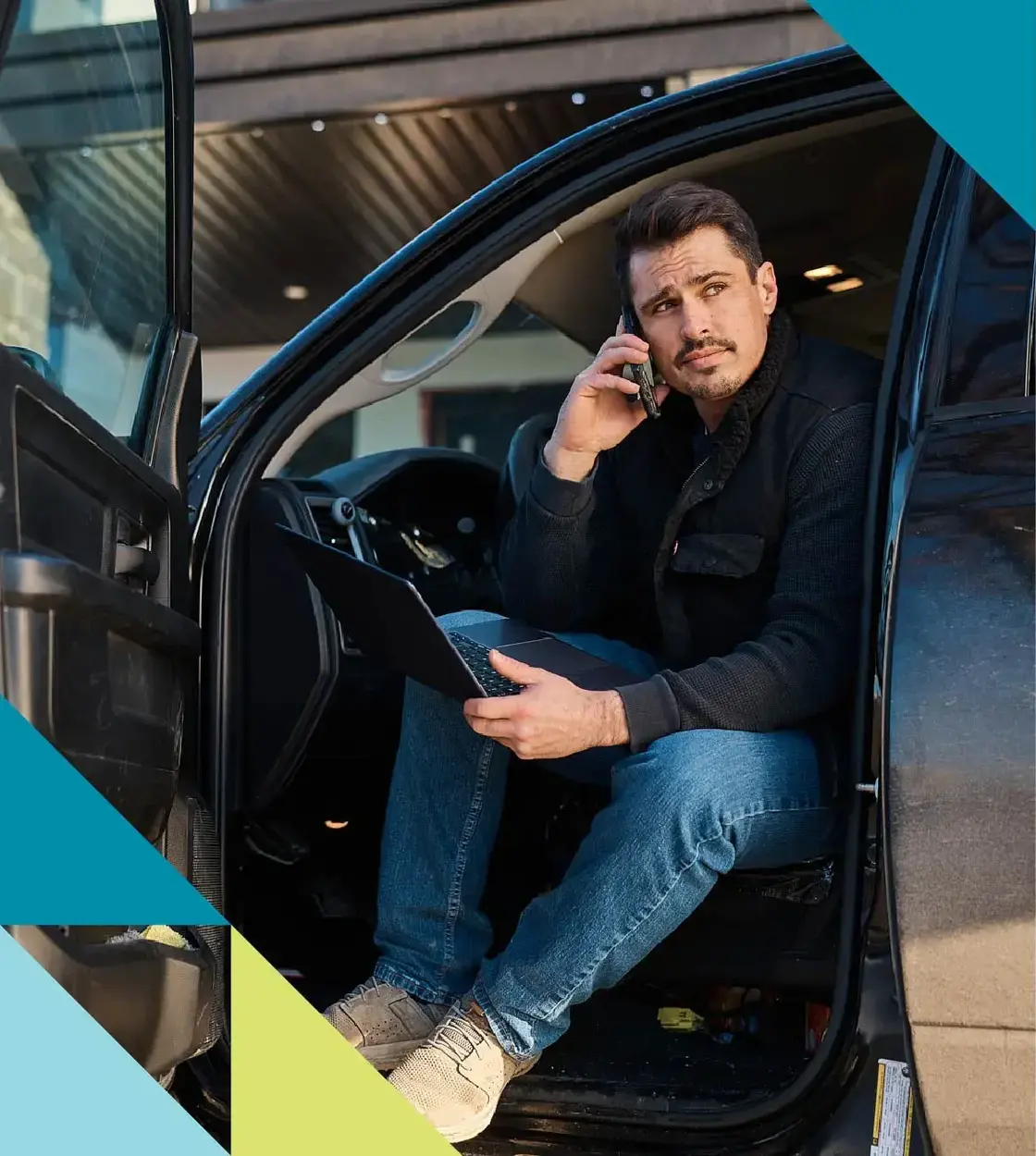 A man with dark hair and goatee leans out his vehicle while taking a call and holding his laptop computer.