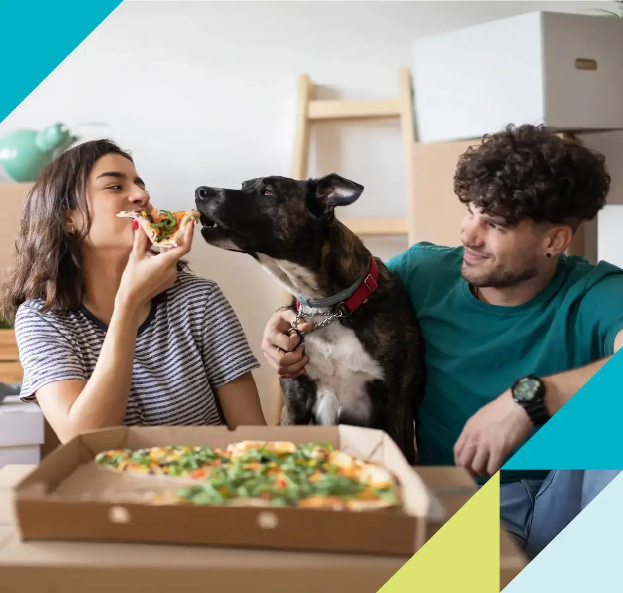 A young couple moving into their new home enjoys a pizza break as their dog tries to snag a slice.
