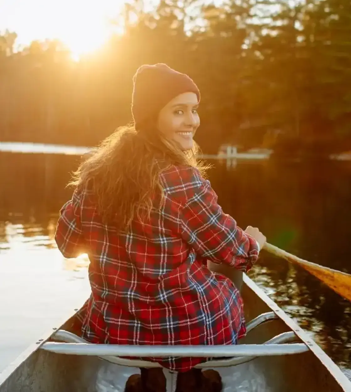 A woman in a red plaid shirt and toque smiles back at the camera as she paddles her canoe on a calm lake.