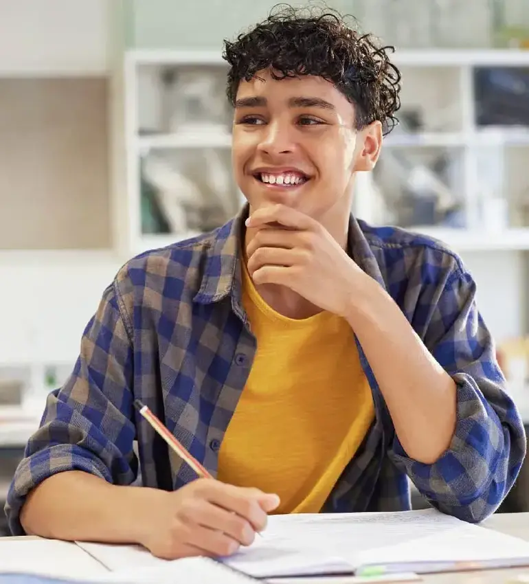 A teenage boy in flannel smiles while taking notes in class.