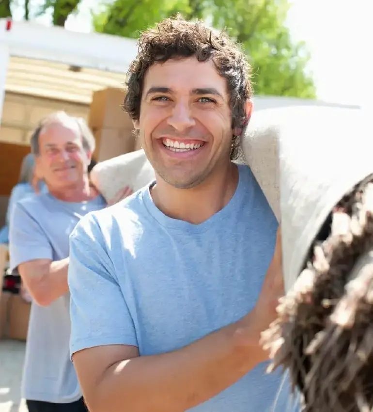 Two male movers in blue t-shirts smile as they haul out a large roll of carpet.