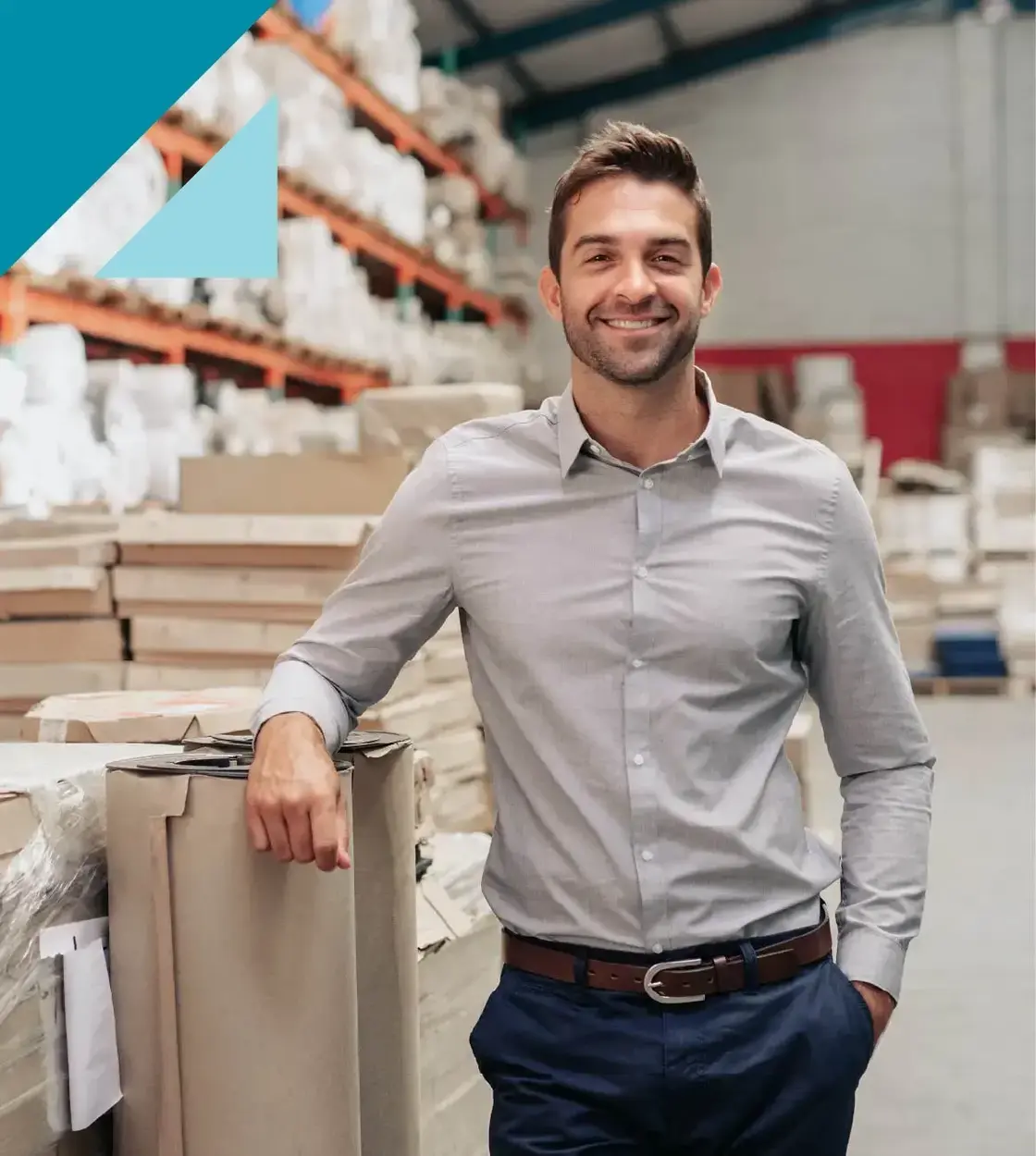 A young man wearing a grey dress shirt smiles at the camera in a warehouse.