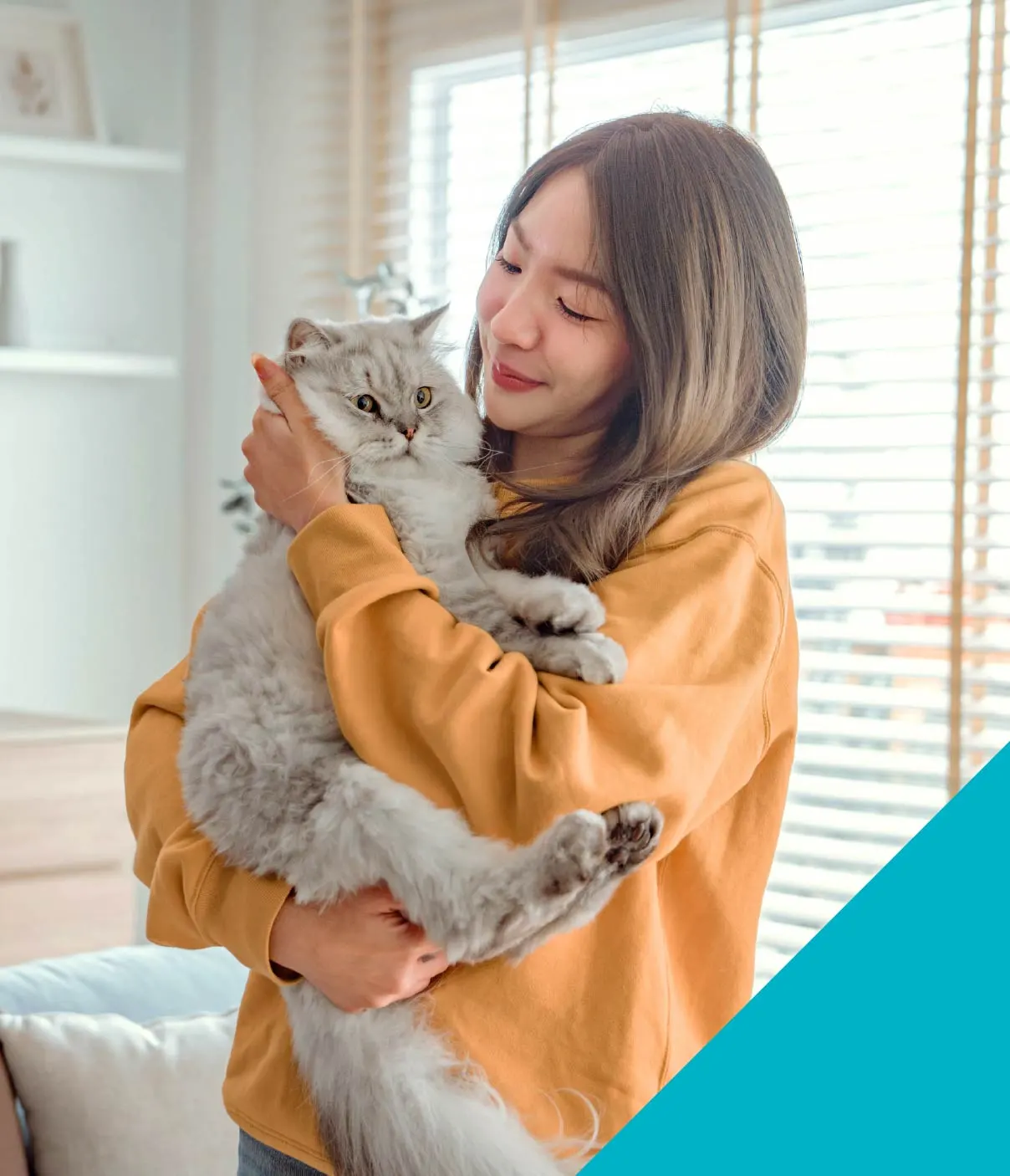 A young woman in an orange sweatshirt smiles at the fluffy grey cat in her arms.