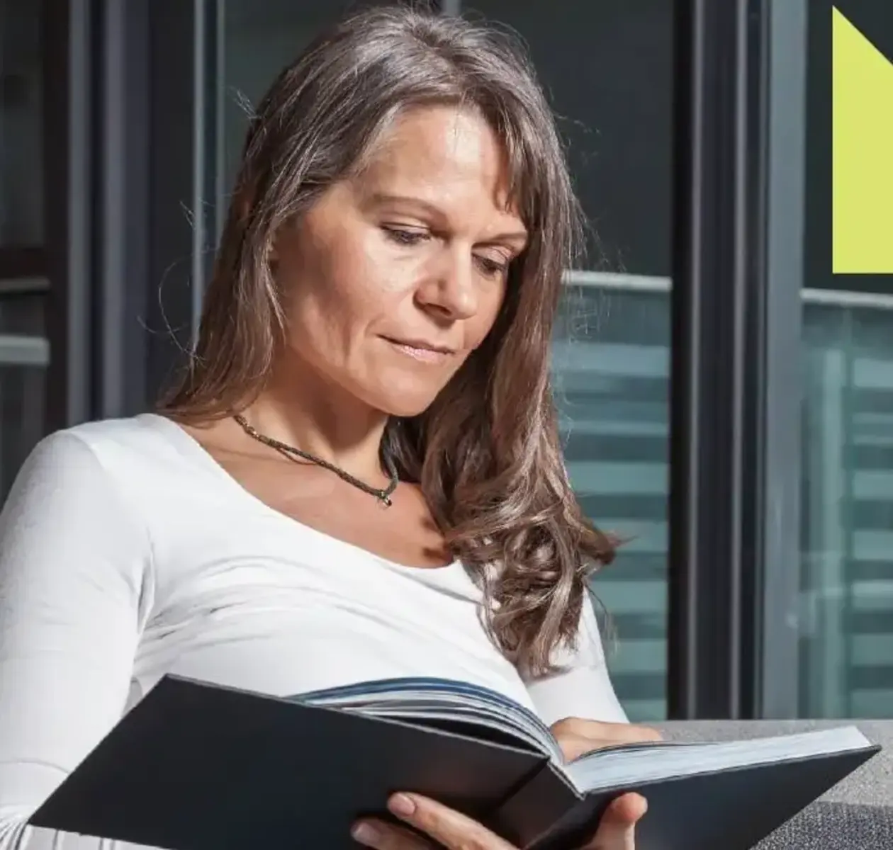A woman with long grey hair relaxes on her couch reading a book.