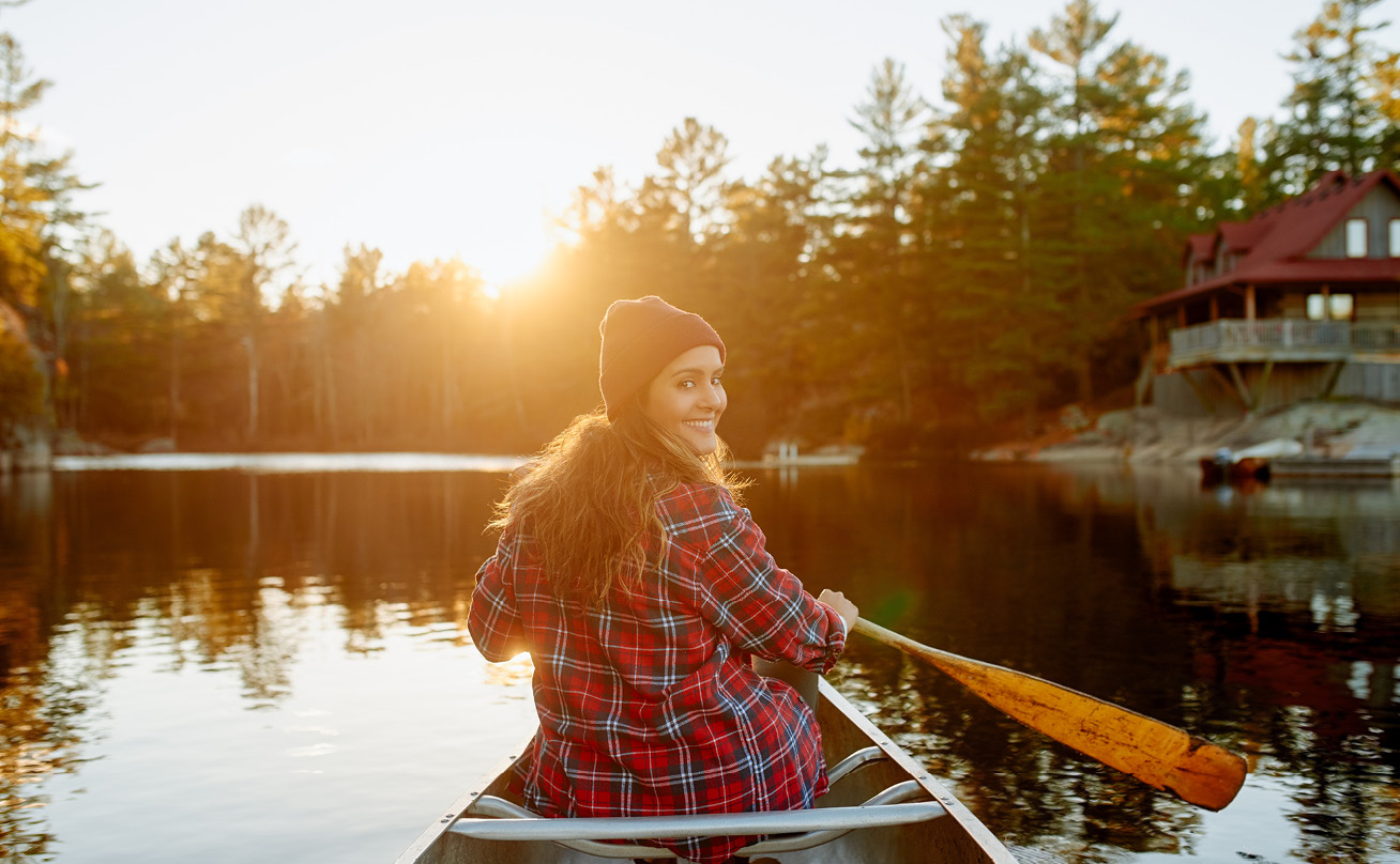A young woman wearing plaid paddles a canoe on a lake at sunset.