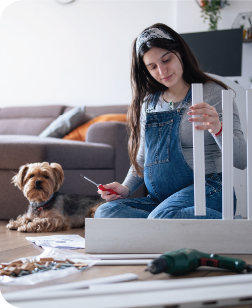 An expecting mother assembles a crib with a screwdriver while her dog watches.