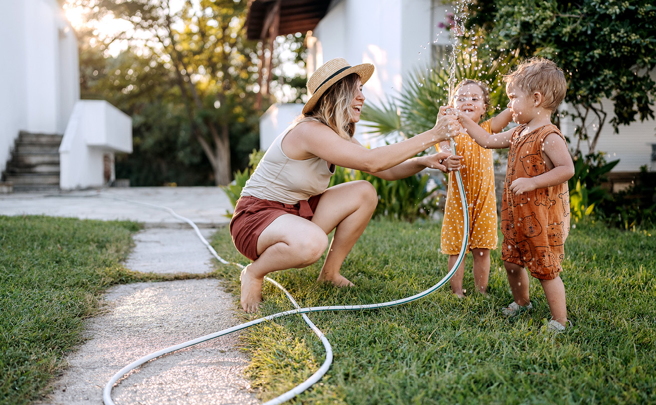 A woman holding a garden hose sprays water next to two young children.
